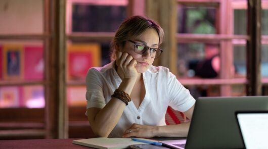 Young woman with blonde hair in a ponytail sitting in front of a laptop and looking unhappy.