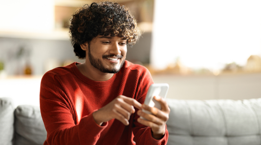 A man wearing a long-sleeved red t-shirt sitting on a couch holding a smartphone and smiling