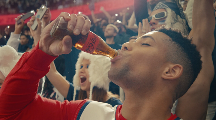 A Coke campaign image showing a young man drinking out of a glass bottle at a basketball game