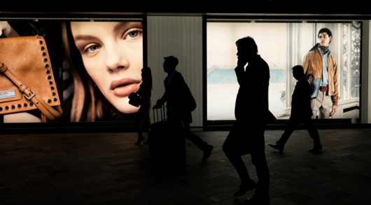 An image of people in shadow walking past a shop with a large-scale close-up image of a woman's face in the window