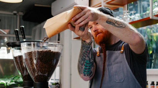 a bearded barista pouring coffee beans into a hopper in an industrial cafe