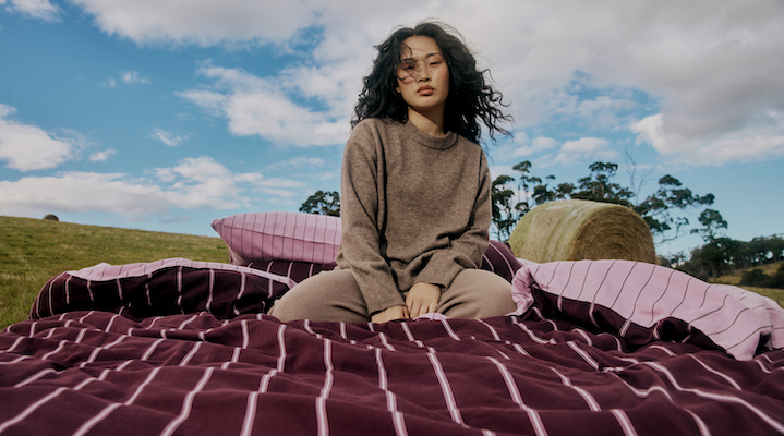 A female model is sitting on homegoods brand Hommey’s Versa Bedding collection on a bed in the middle of a field.
