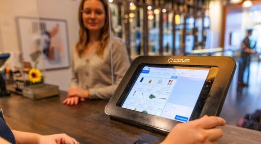 A blond woman wearing glasses is standing in front of a desk and a sales associate using an Ipad.
