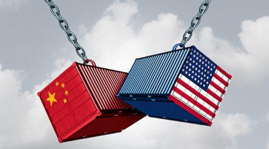 One block with the US flag and one block with the Chinese flag are suspended on chains, hitting one another, set against a cloudy blue sky.