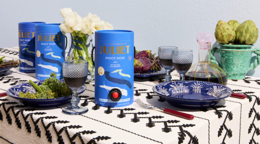 A container of Juliet Wine’s pinot noir is set on a dining room table with a printed tablecloth, set against a light grey background.