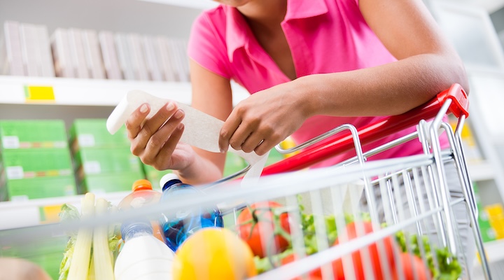 Woman checking a long grocery receipt leaning to a full shopping cart at store.