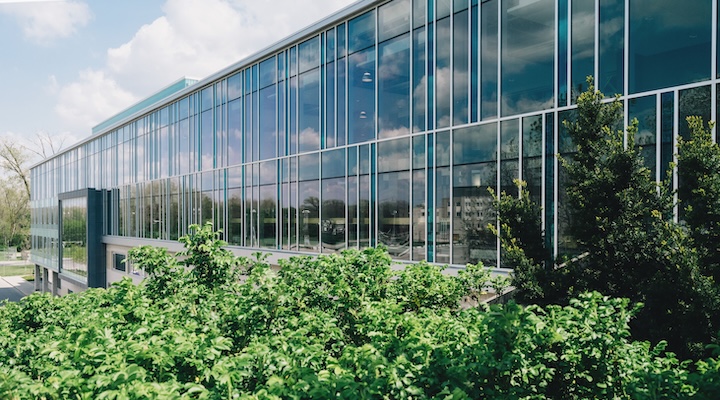 Contemporary building facade with glass panels reflecting nature, surrounded by lush greenery.