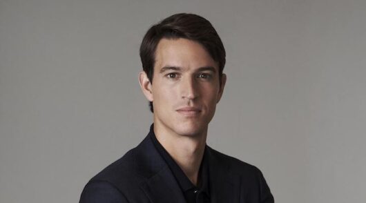 LVMH heir Alexandre Arnault standing in front of a grey background in a black shirt.