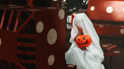 A person dressed as a ghost holding a jack-o'-lantern, sitting on a red and white bench.