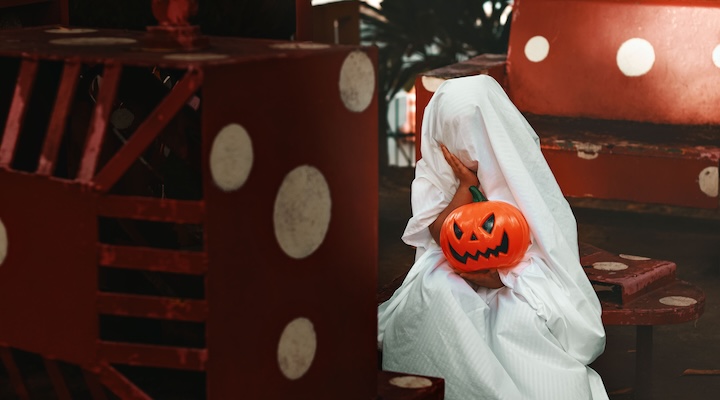 A person dressed as a ghost holding a jack-o'-lantern, sitting on a red and white bench.
